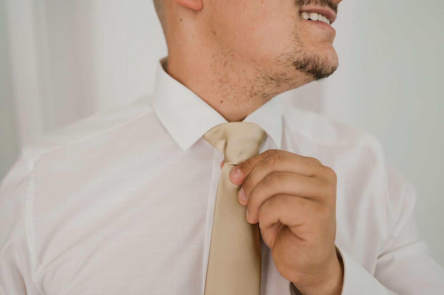 Groom adjusts champagne tie while wearing crisp white dress shirt.