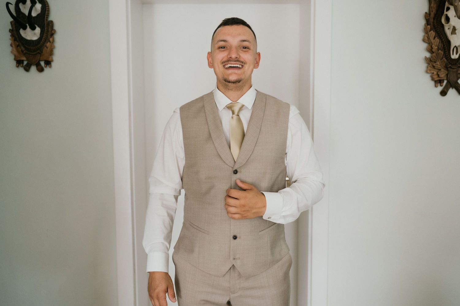 A groomsman wearing a beige vest and white dress shirt smiles joyfully in a minimalist interior setting.