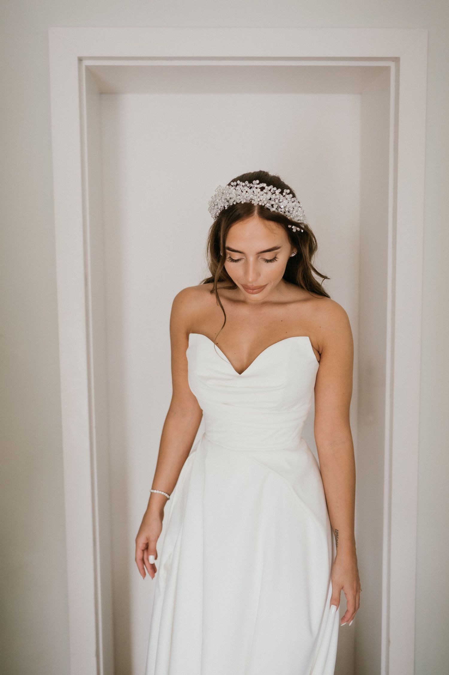 A bride wearing a strapless white gown and crystal headpiece looks down gracefully.