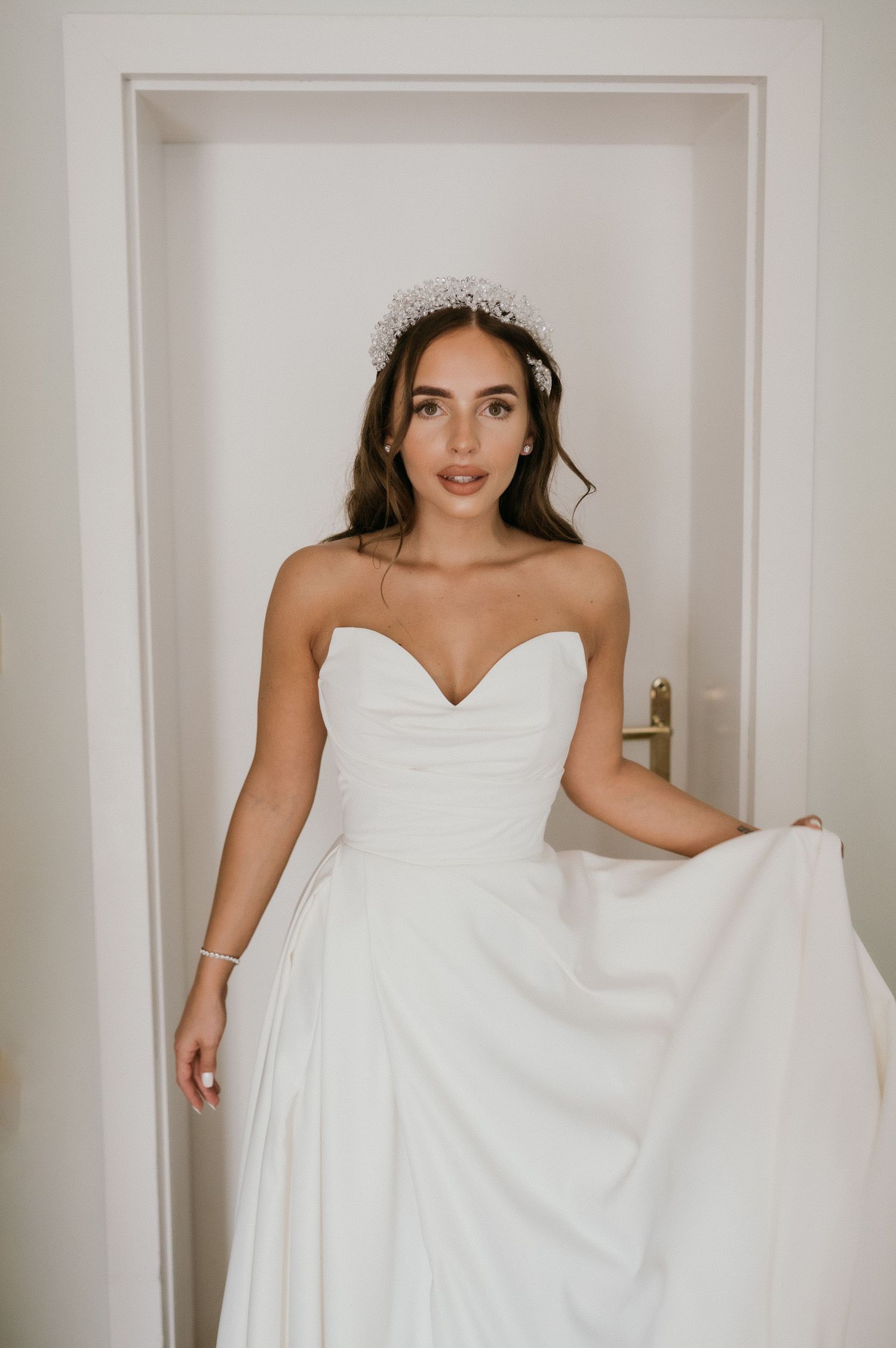 A bride in a strapless white wedding gown and crystal crown poses elegantly in a doorway.