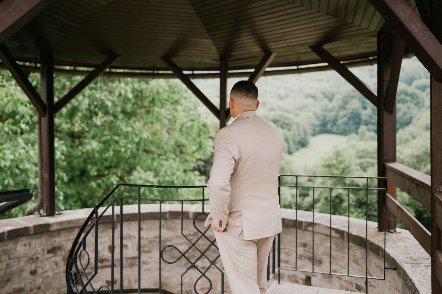 A person in a light-colored suit stands on a covered balcony overlooking a scenic view.