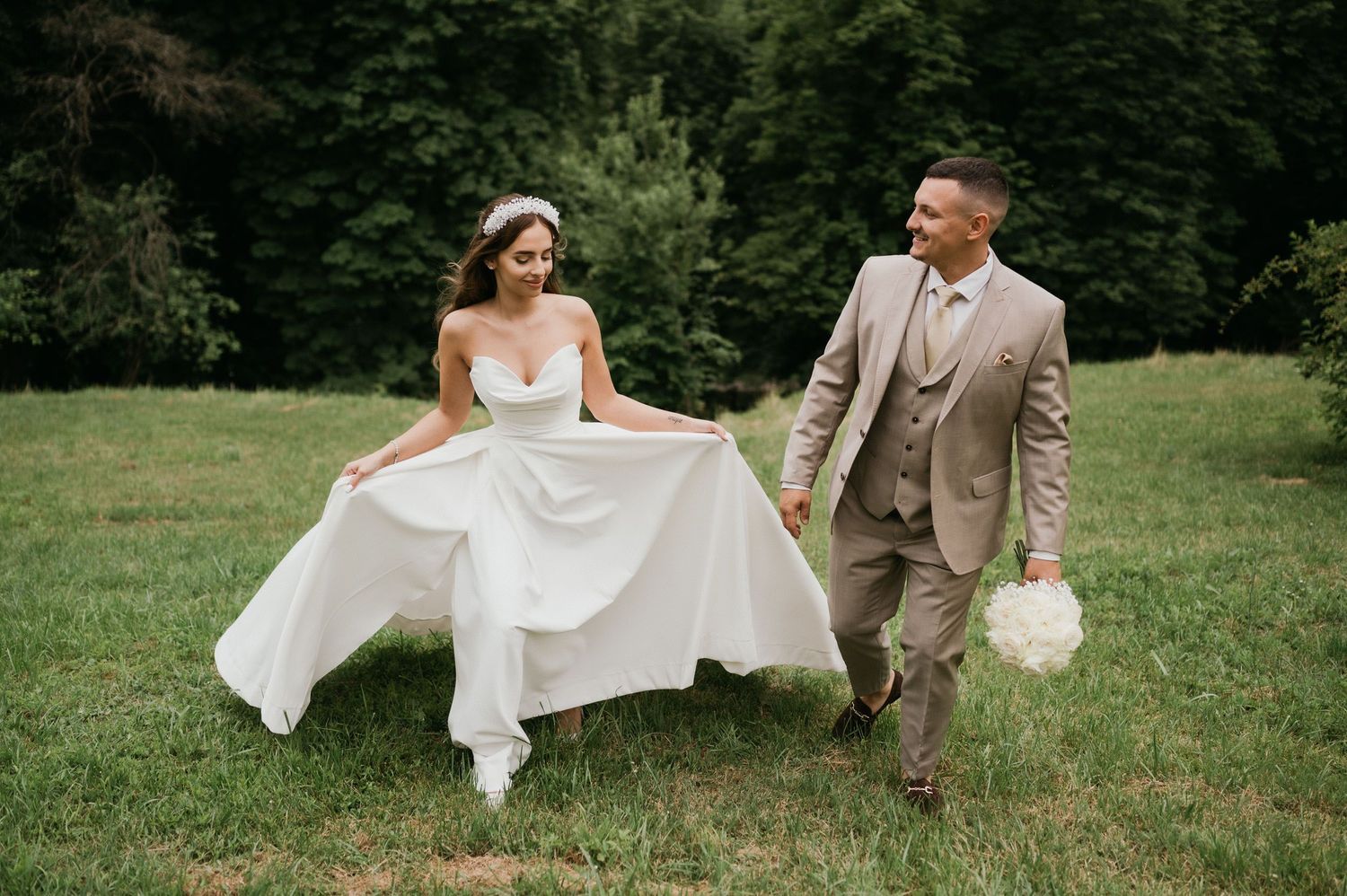 Newlyweds walk through a grassy field as bride playfully holds her flowing white dress.