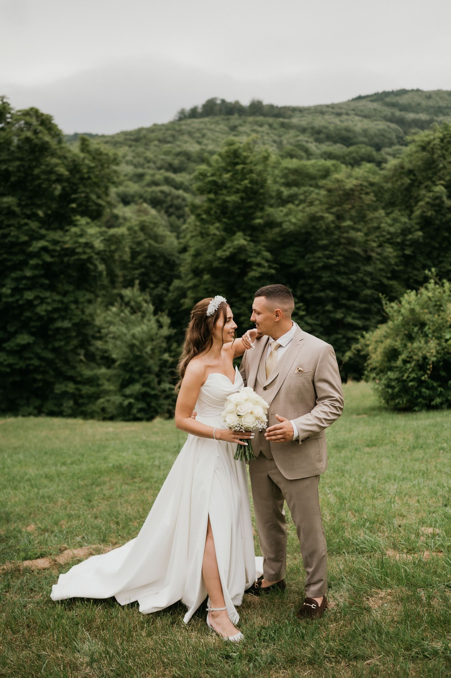 Newlyweds pose together on a hillside with white bouquet and elegant wedding attire.