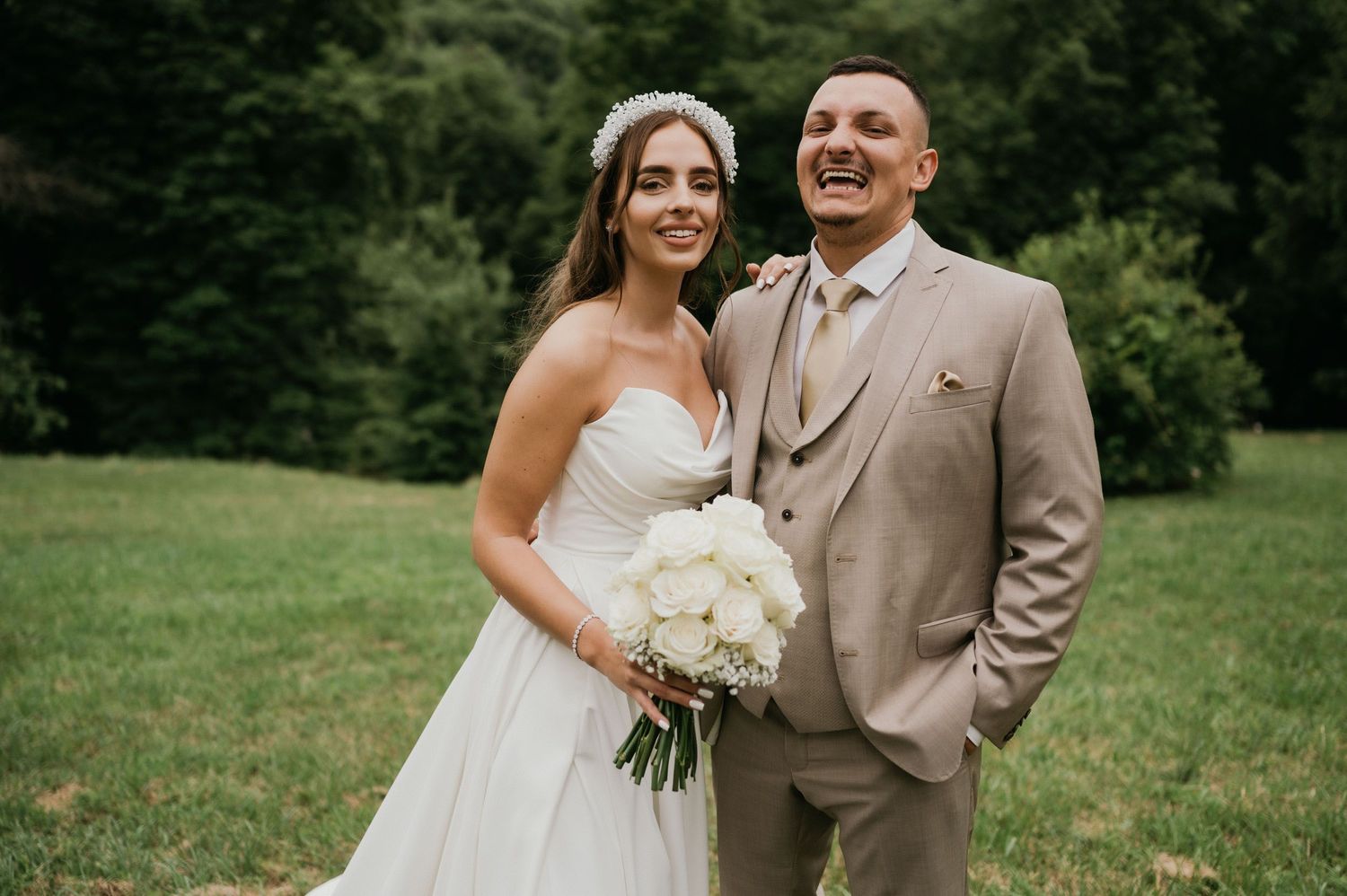 A couple in wedding attire smiles while standing in a lush green outdoor setting.
