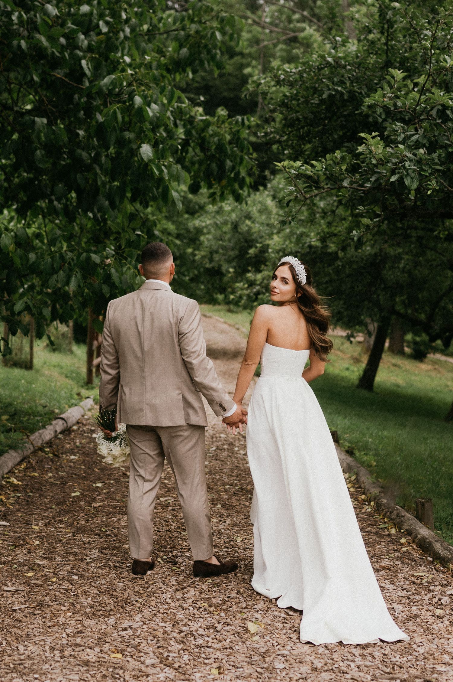 Bride and groom walk hand in hand down a tree-lined path on their wedding day.