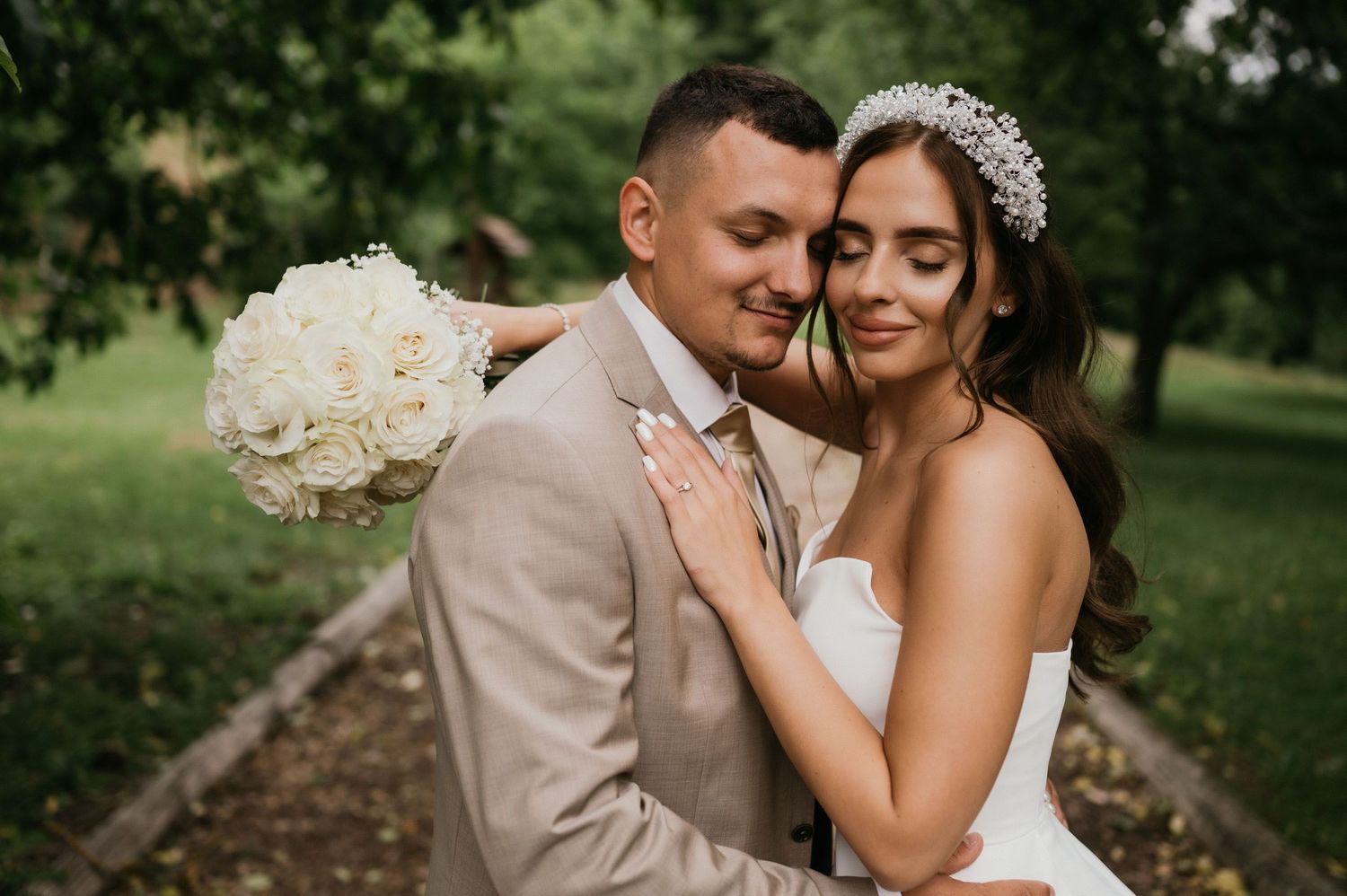 A tender wedding moment captured on a garden path with white floral bouquet and beige suit.