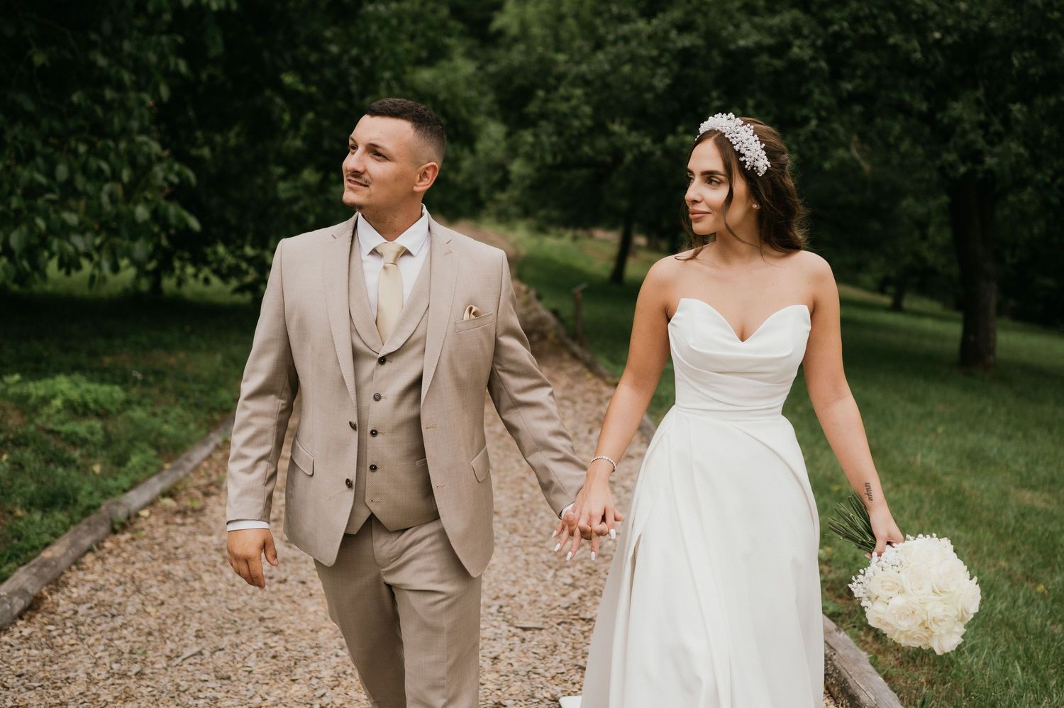 A romantic wedding couple walks hand-in-hand down a gravel path surrounded by lush greenery.