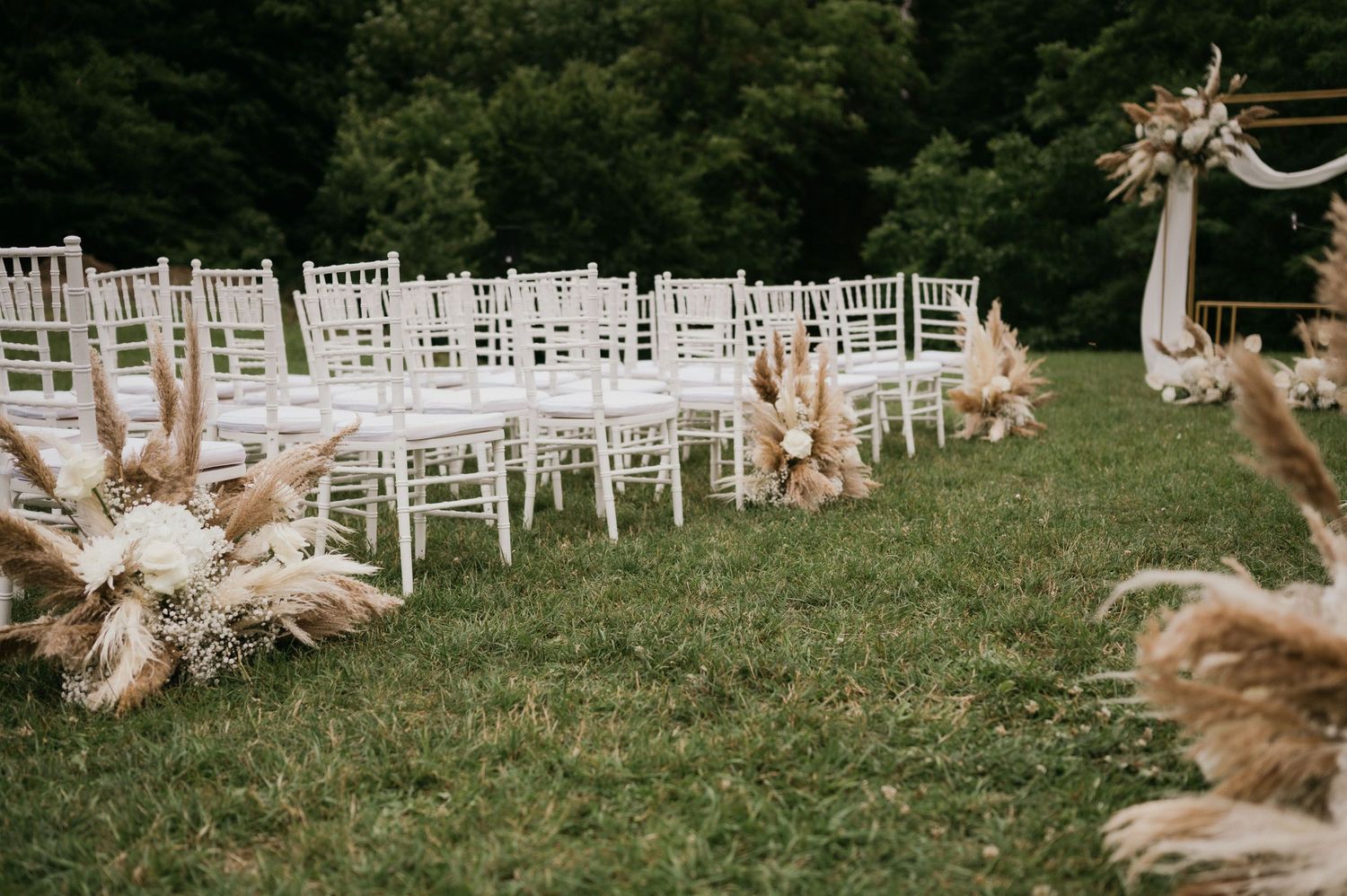 White wedding chairs line a grass aisle decorated with dried pampas grass arrangements.