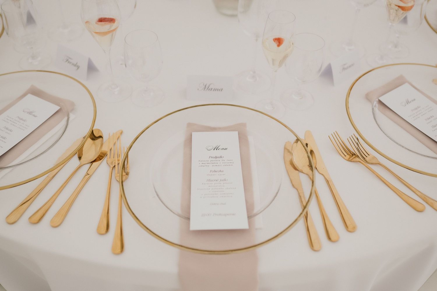 Elegant place setting with gold cutlery, charger plate and menu card on white tablecloth at wedding reception.