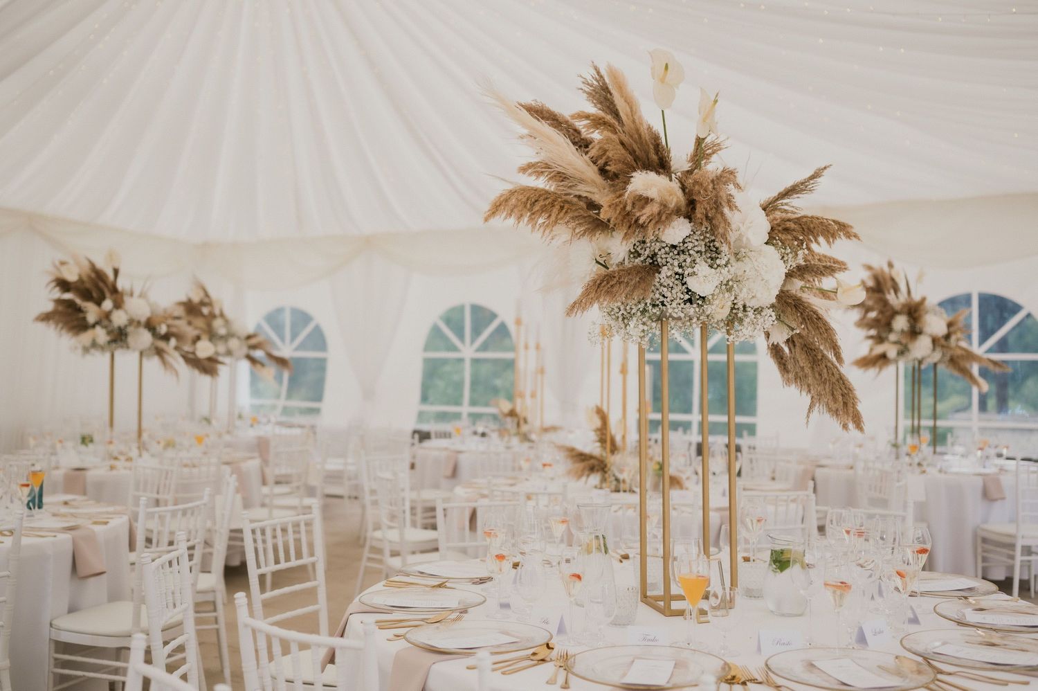 Romantic wedding reception setup in white tent with pampas grass centerpieces and chiavari chairs.