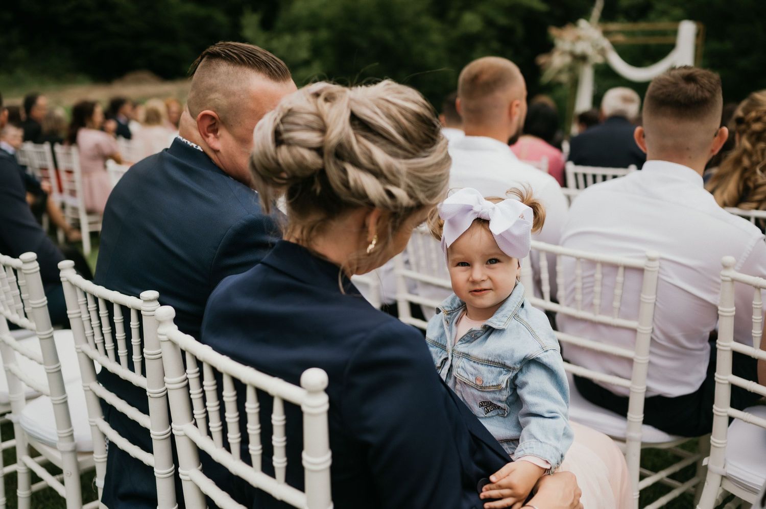 Wedding ceremony guests seated in white chairs watching outdoor proceedings.