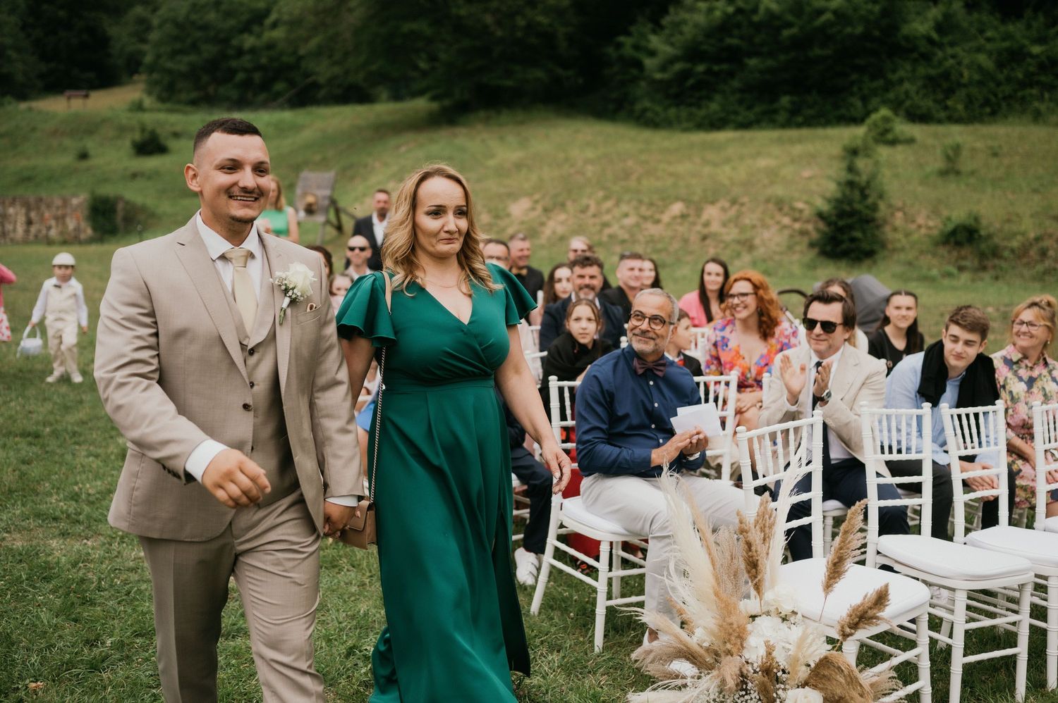 Wedding attendants walk down an outdoor aisle lined with white chairs and pampas grass decor in a grassy field.