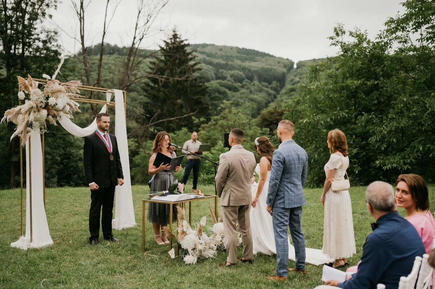 Outdoor wedding ceremony with floral arch and mountains in background as guests gather on lawn.