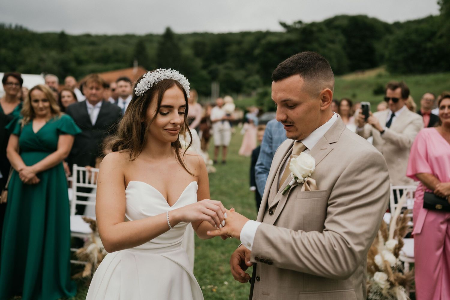 Outdoor wedding ceremony with couple exchanging rings surrounded by guests in formal attire.