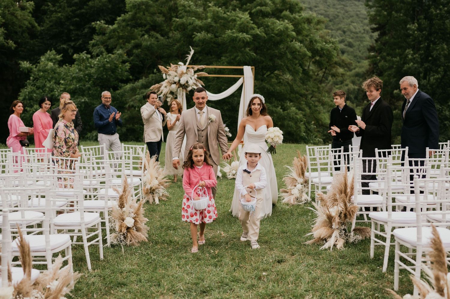 Wedding party walking down aisle between white chairs as guests celebrate with raised hands.