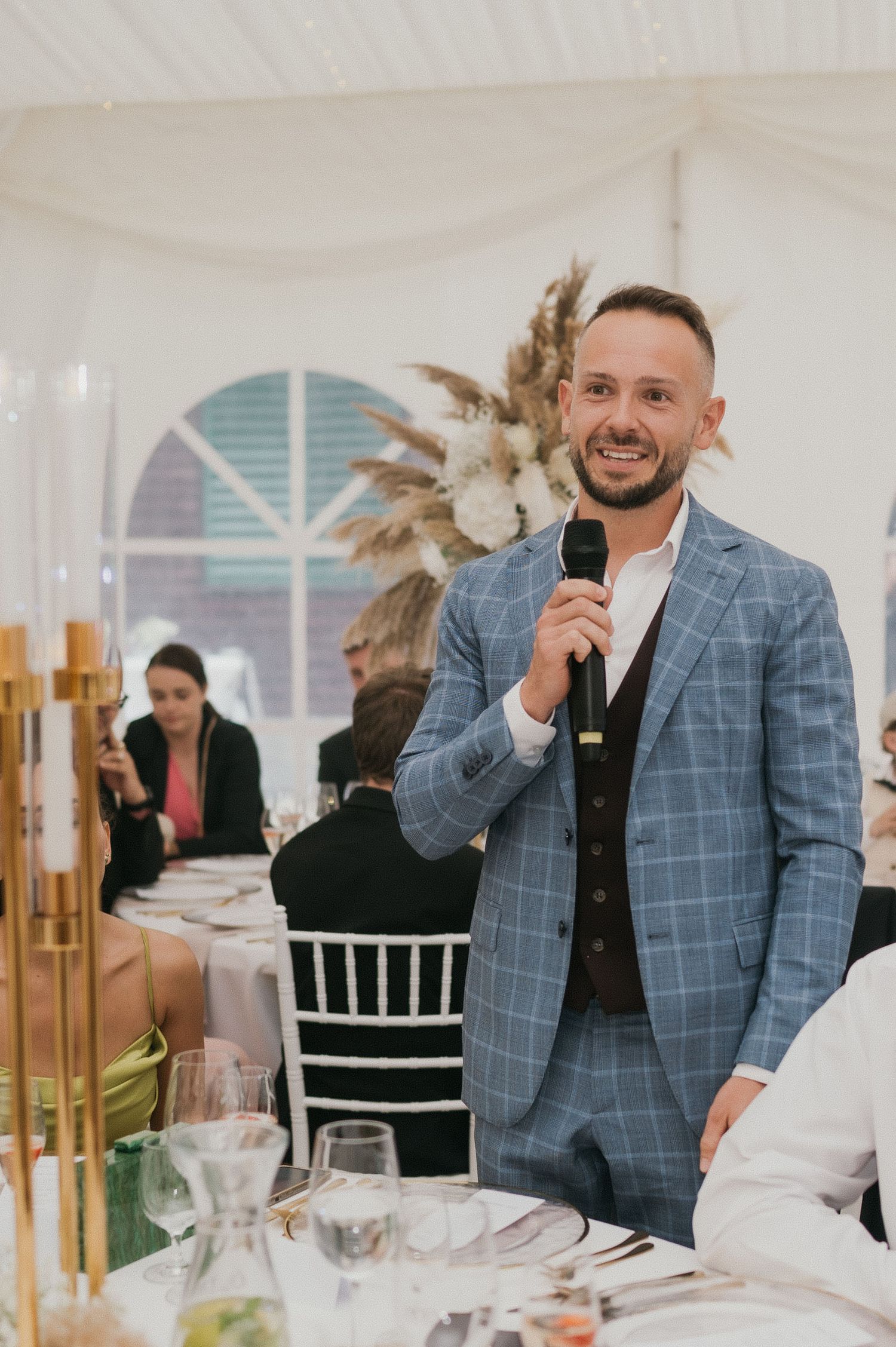 Speaker in blue checkered suit gives speech at elegant wedding reception with floral decorations.