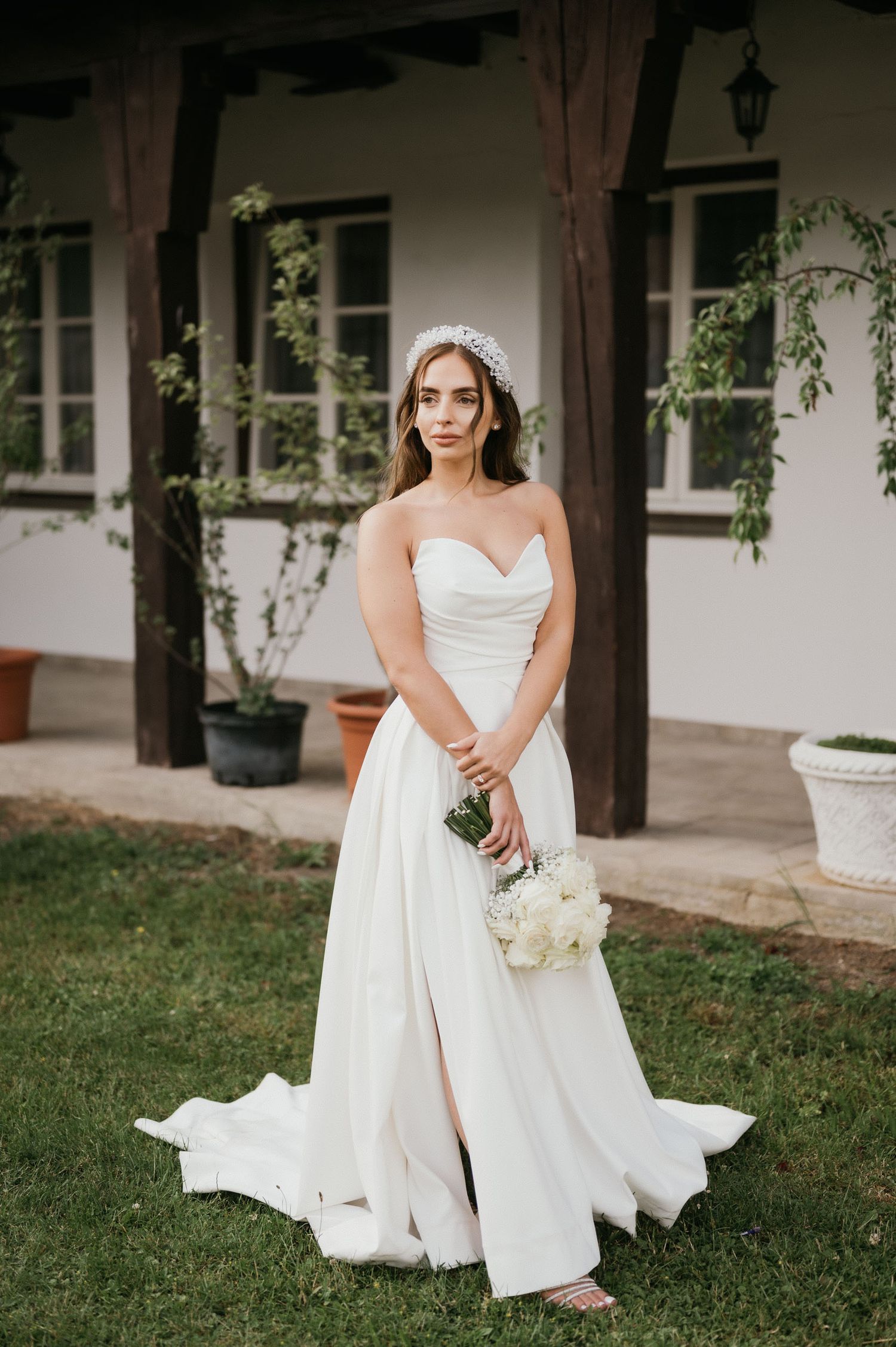 A bride in a strapless wedding gown holds a white rose bouquet against a rustic porch backdrop.