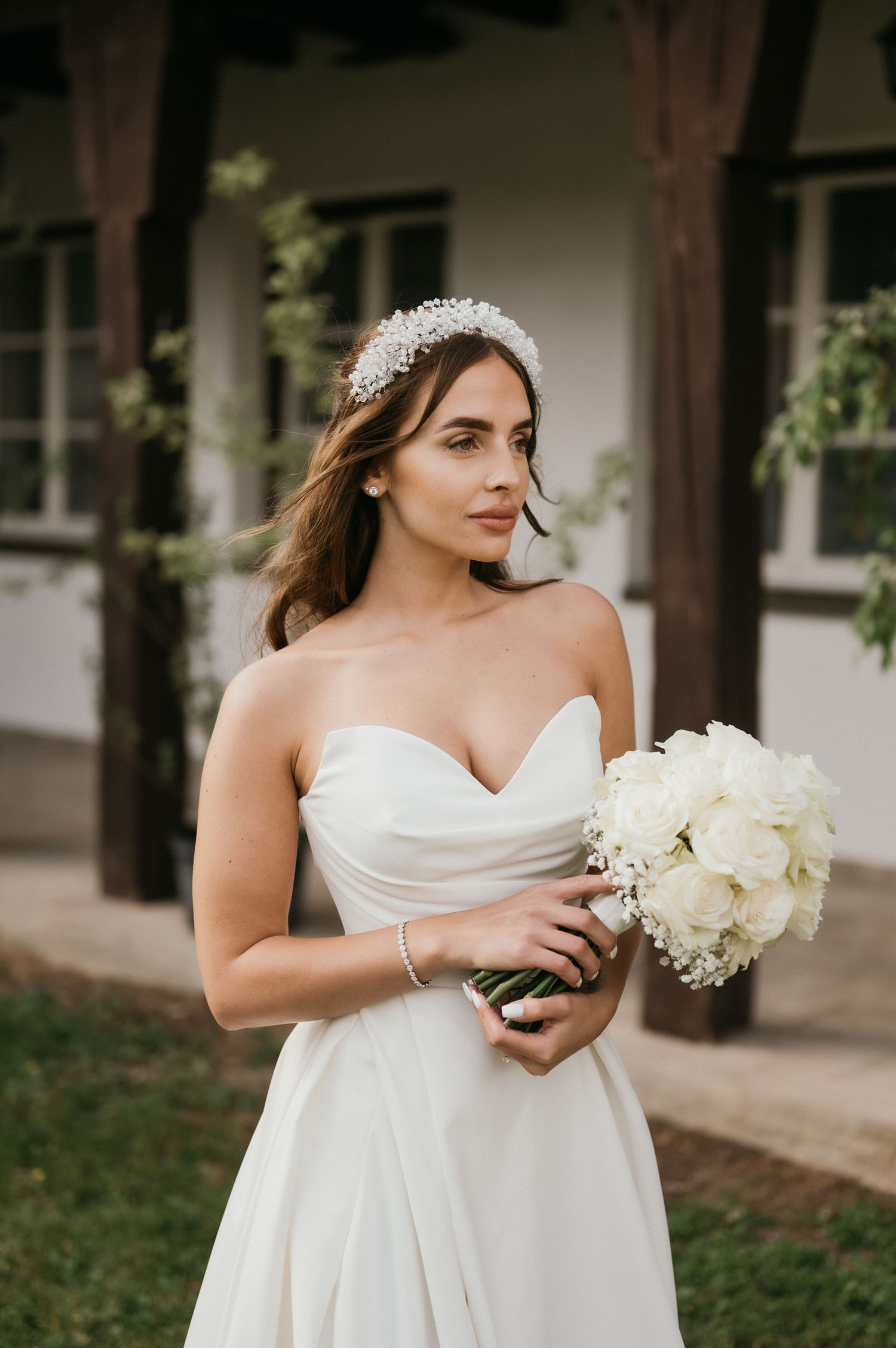 A bride wearing a crystal headpiece holds a bouquet of white roses against dark wooden posts.