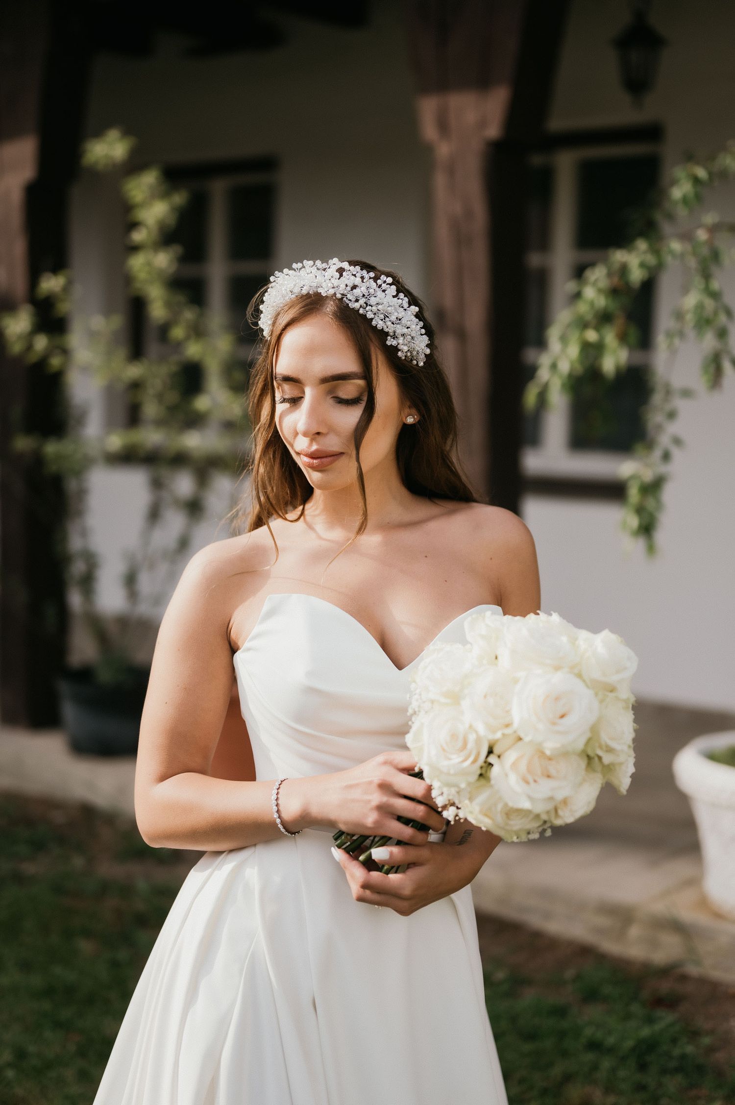 Bride in white strapless gown with delicate lace headpiece holds elegant white rose bouquet against rustic backdrop.
