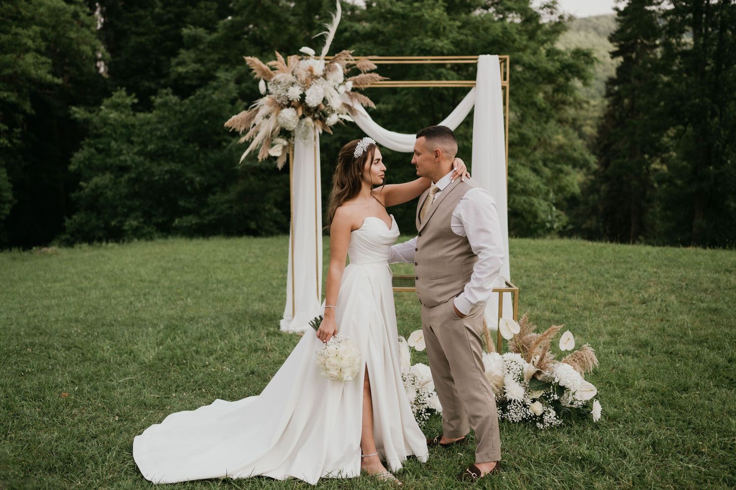 Wedding ceremony setup with white arch decorated with pampas grass and florals on green lawn.