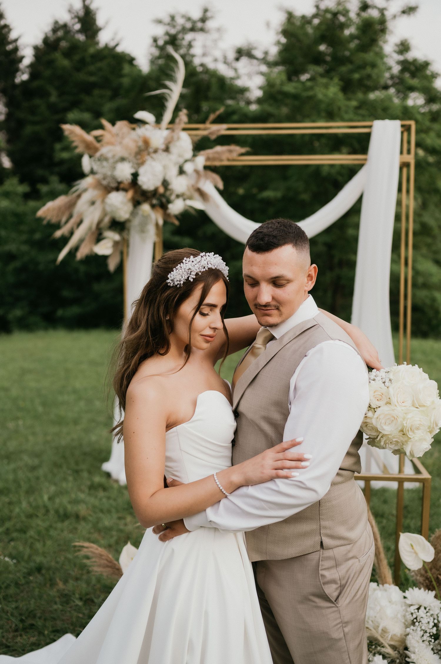 Newlyweds embrace in front of a gold arch decorated with pampas grass and white flowers during an outdoor ceremony.