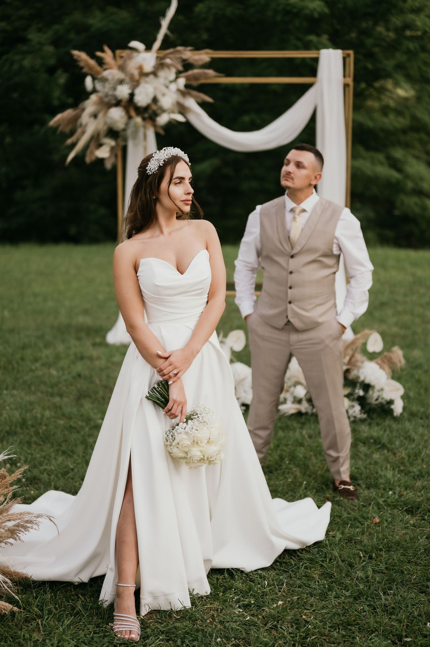 Wedding couple poses elegantly under floral arch with bride in split dress and groom in tan suit.