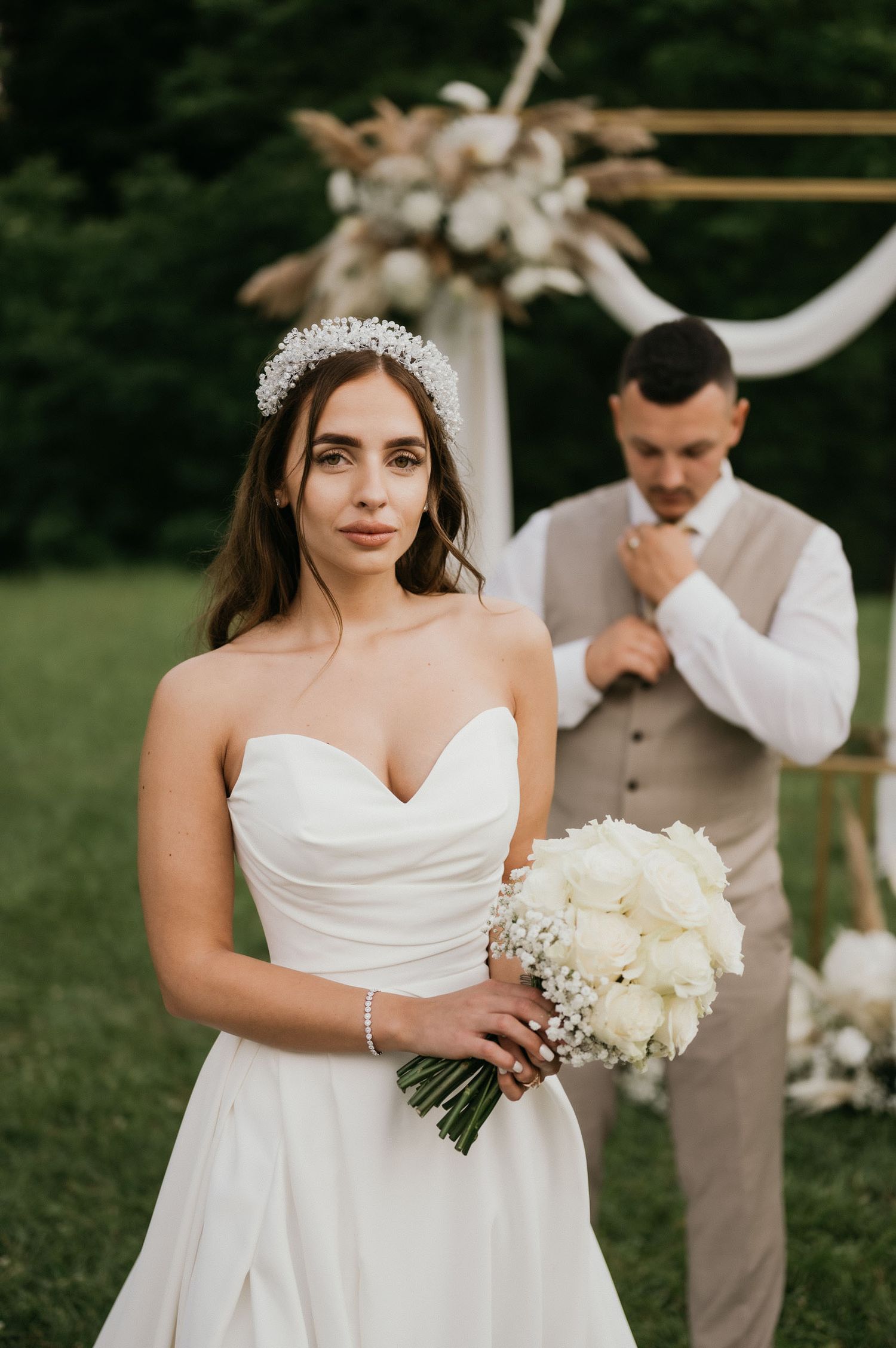 Bride in strapless gown holds white bouquet while groom adjusts tie during outdoor wedding portraits.