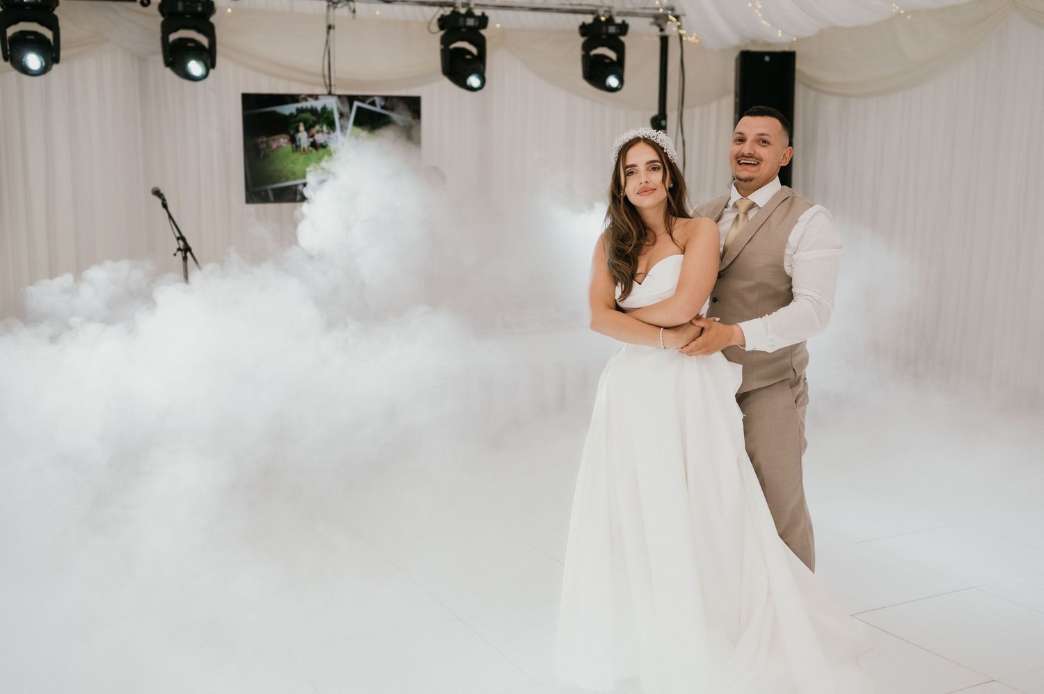 Newlyweds dancing in misty atmosphere with stage lighting at indoor reception venue.