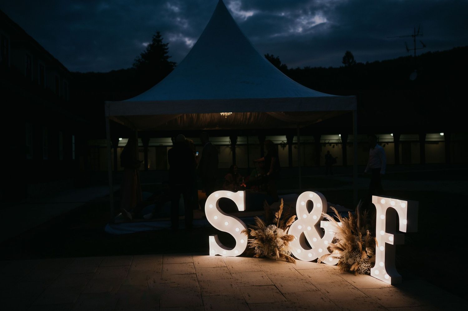 Illuminated letters S&F glow against dark sky at outdoor wedding venue.