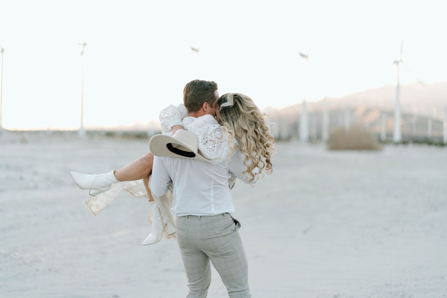 a bride in a white lace dress and groom in relaxed wedding attire pose in front of the windmills in palm springs while spinning