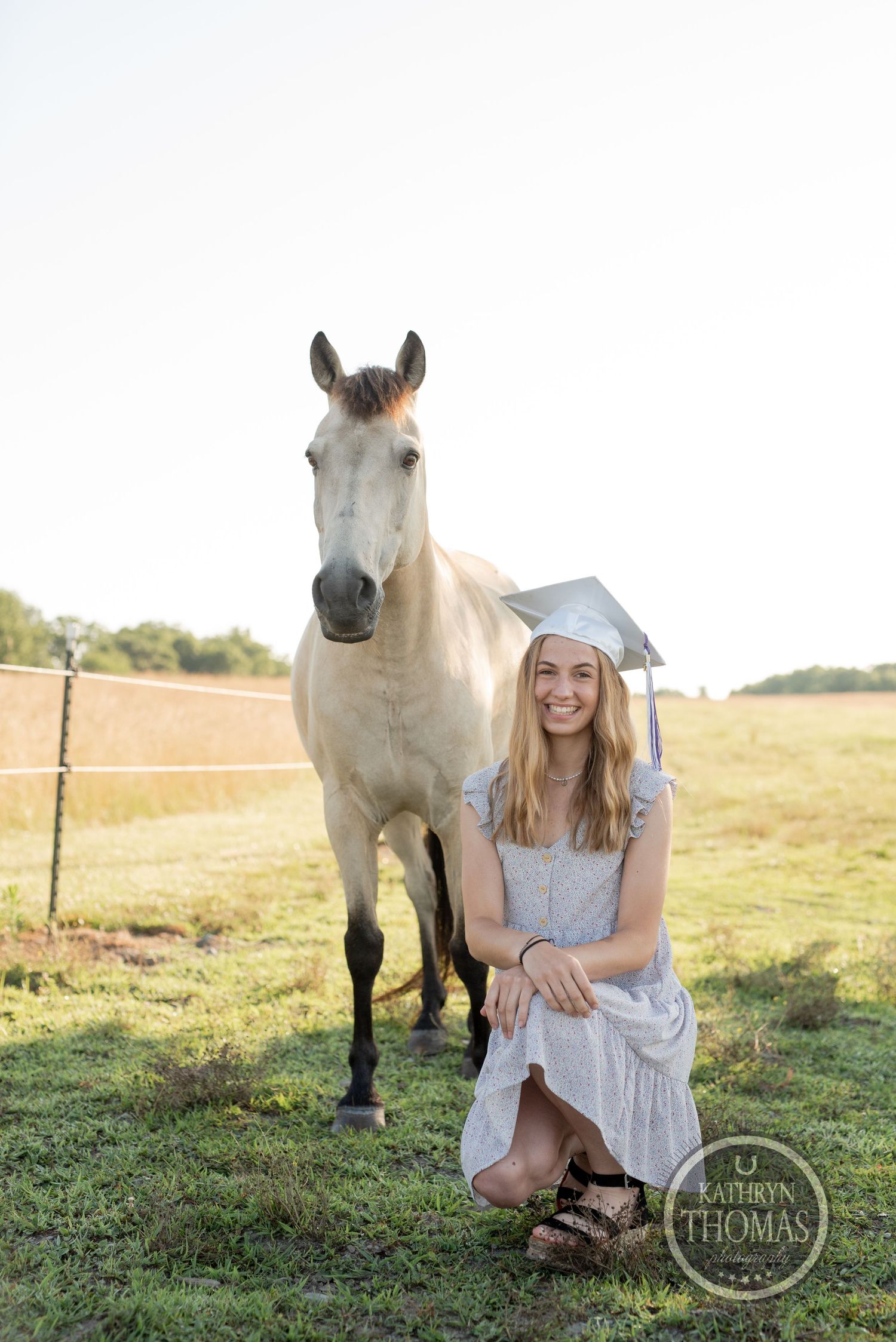 A white horse stands beside someone in a light dress holding papers in a sunny field.