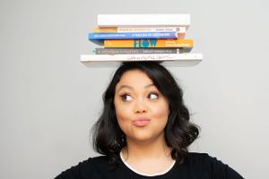 Woman in black outfit playfully posing with stack of books against gray background.