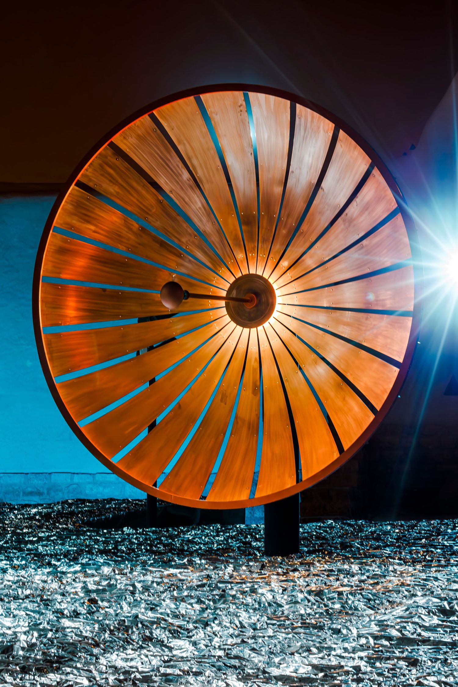 Backlit wooden circular sculpture with radiating slats creates dramatic lighting pattern at night.