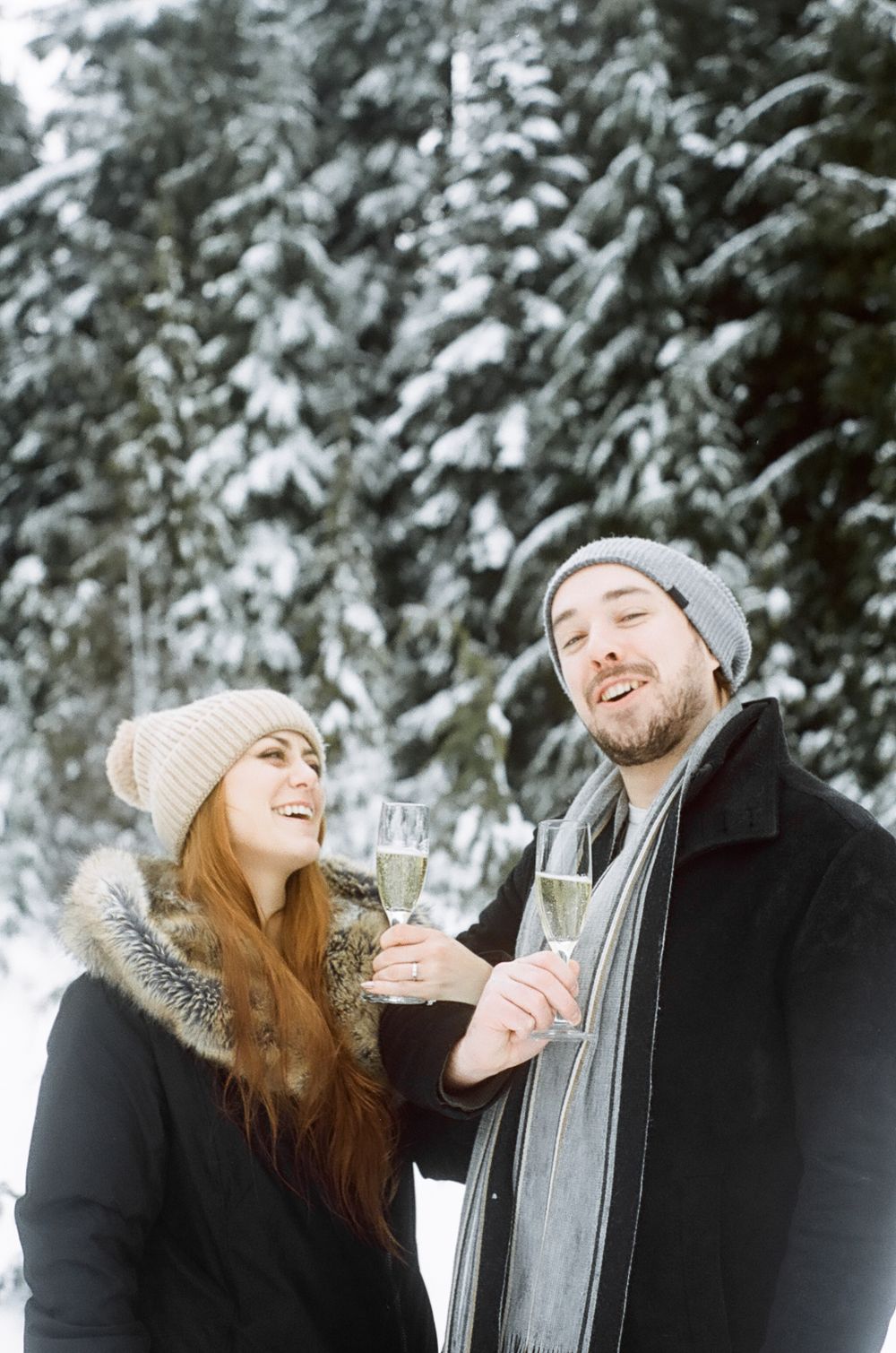Cheerful couple celebrating with champagne glasses in snow-covered forest setting.