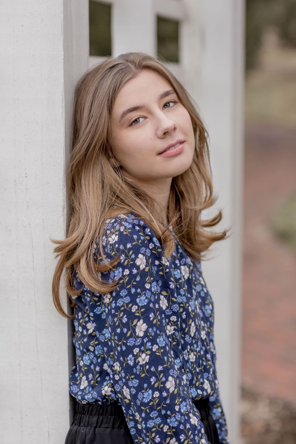 Young woman in blue floral blouse leaning against white fence post outdoors.