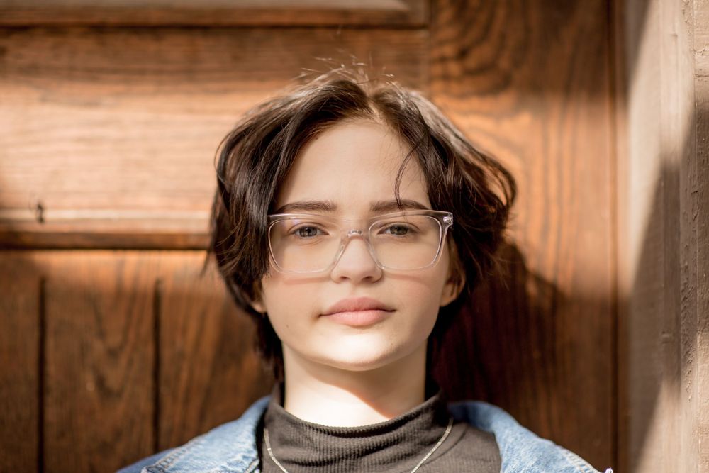 Young person with short dark hair and clear glasses leaning against a wooden door.