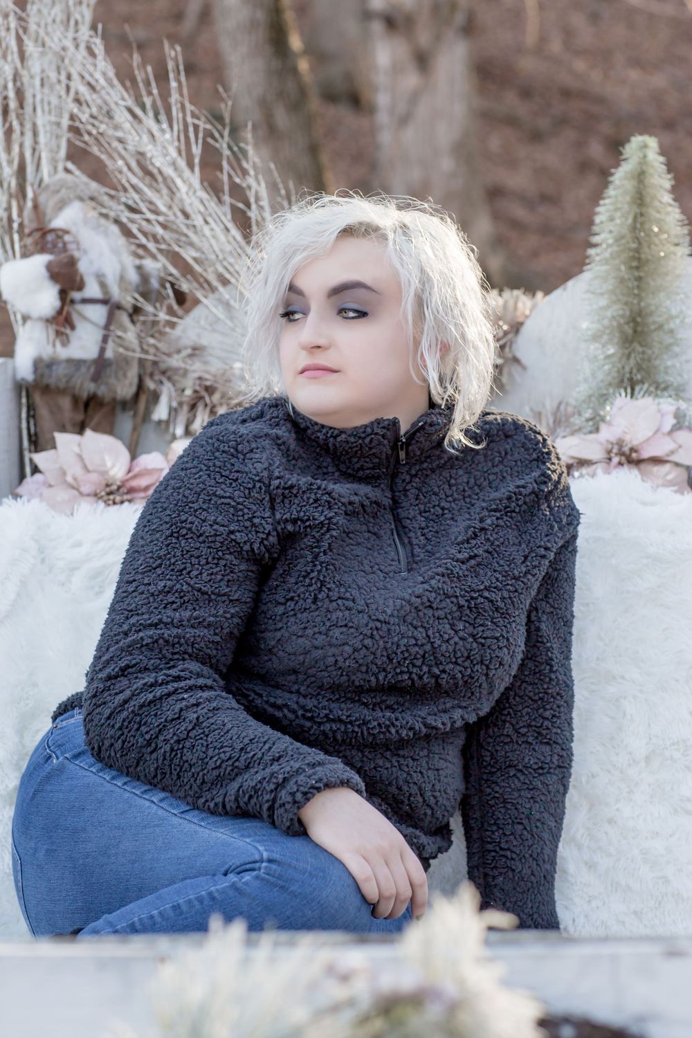 Blonde teen in gray fleece pullover and jeans sitting outdoors among winter decorations.