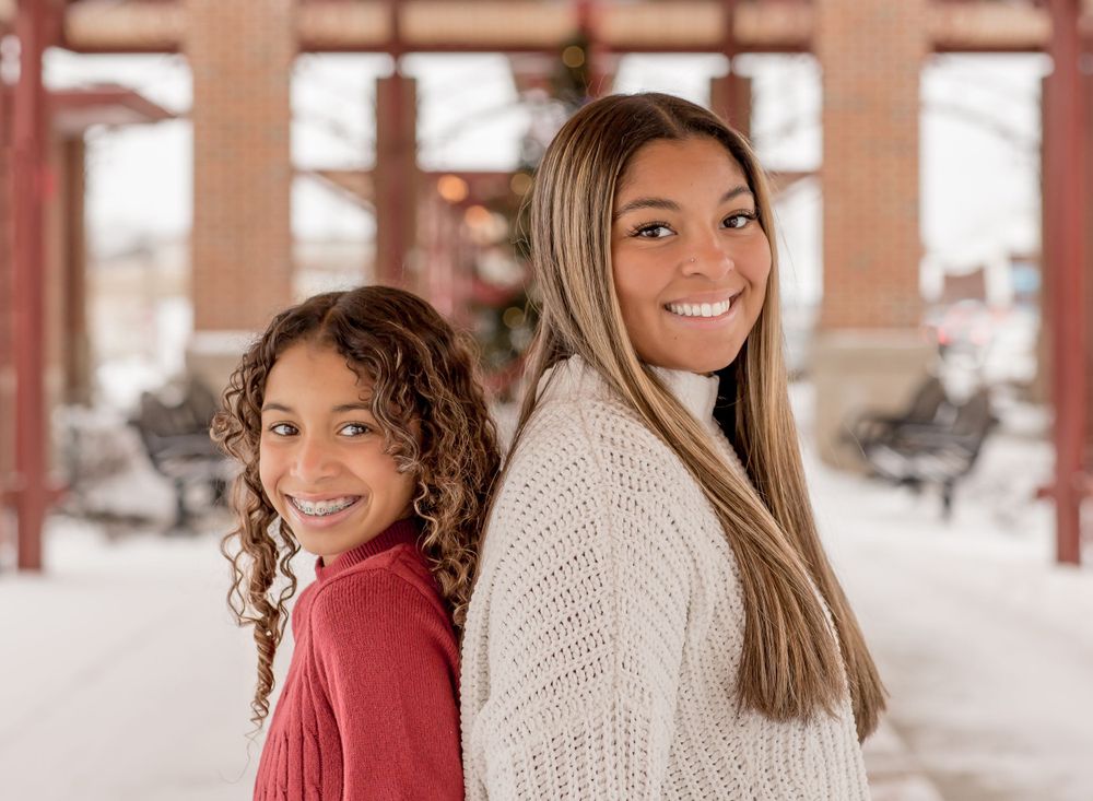 Two smiling girls posing back-to-back in winter sweaters at snowy outdoor location.