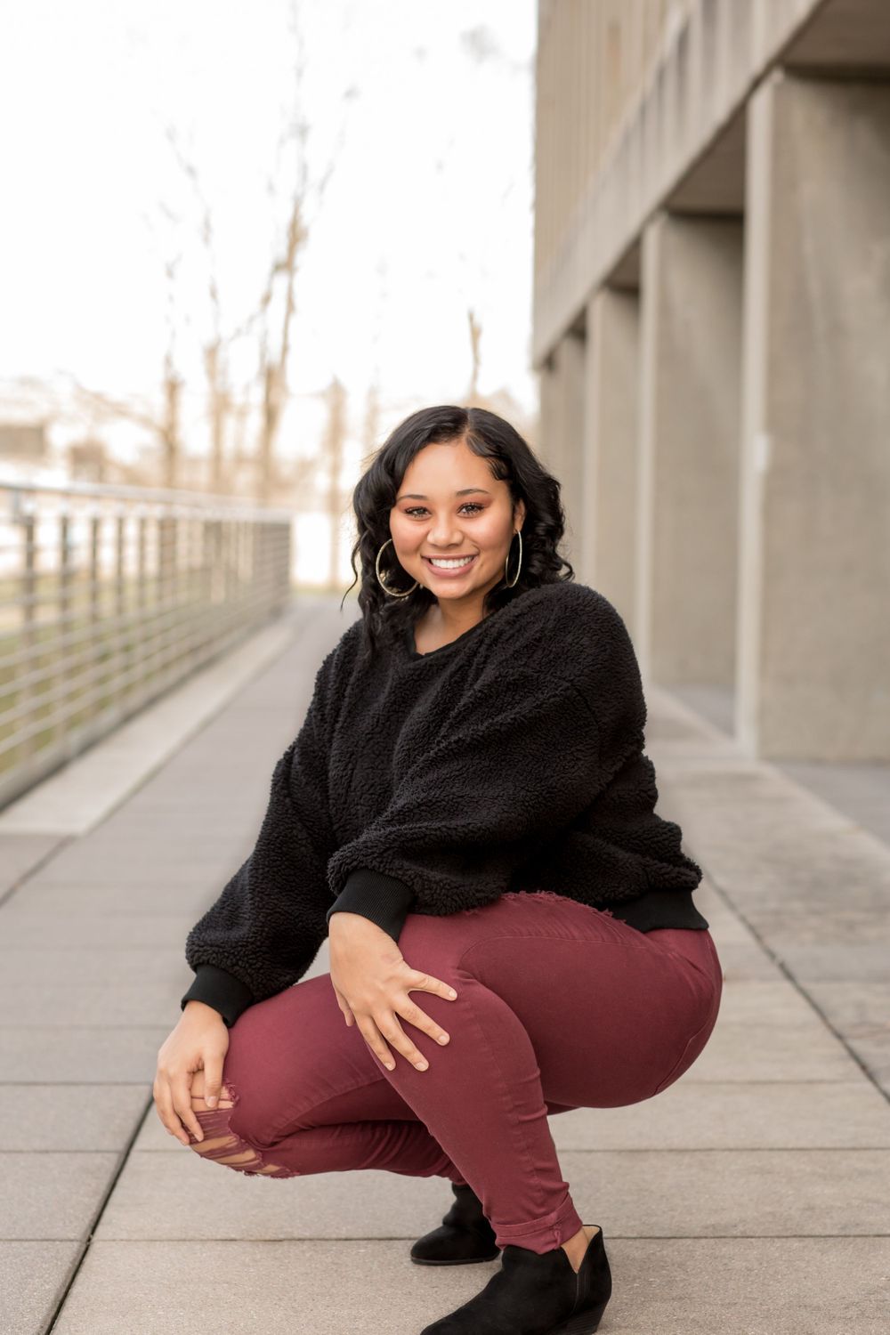 Smiling young woman in black sweater and maroon pants crouching outdoors.