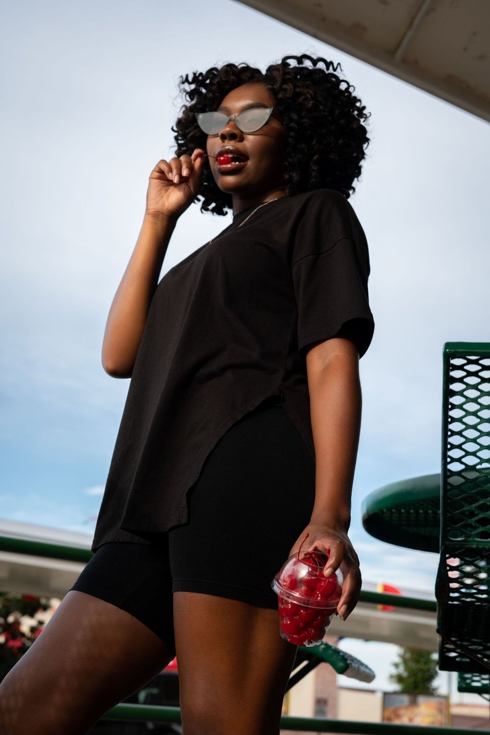 Urban fashion portrait of someone in black outfit holding red drink with curly hair and glasses.