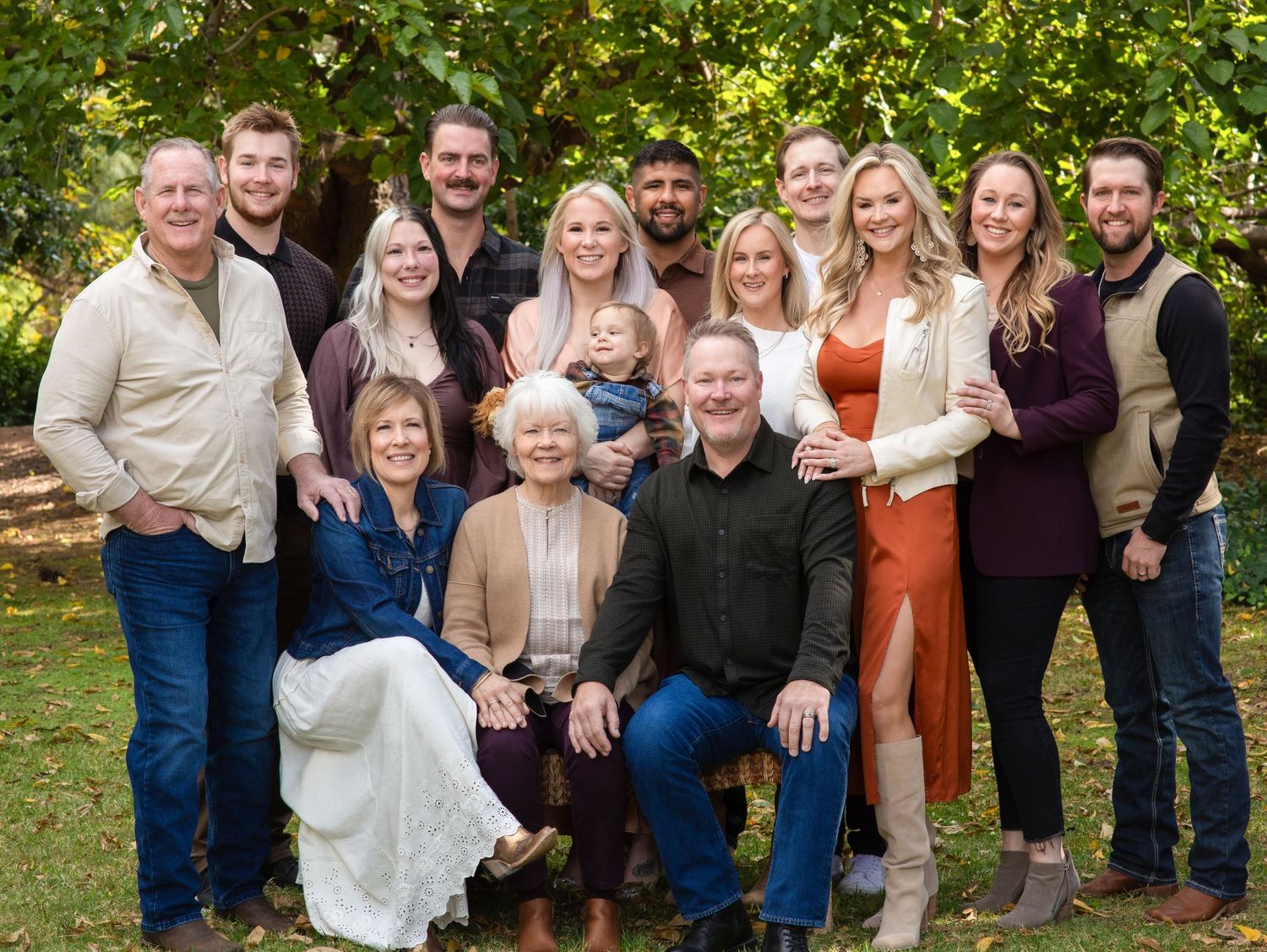 A large family portrait taken outdoors with multiple generations gathered together in casual attire against greenery.