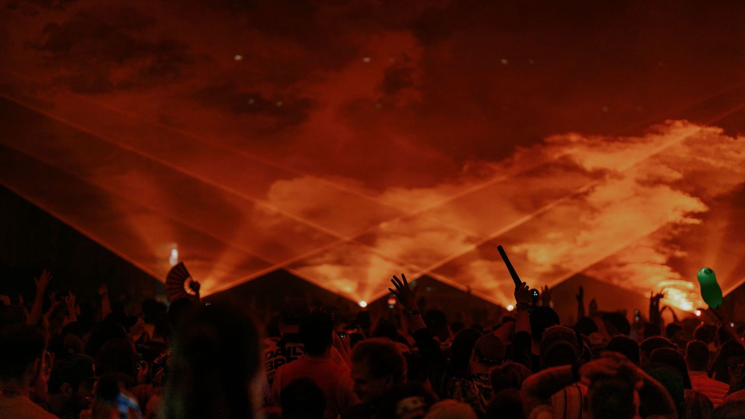 Concert crowd silhouettes against dramatic orange and red sky with light beams cutting through smoke.