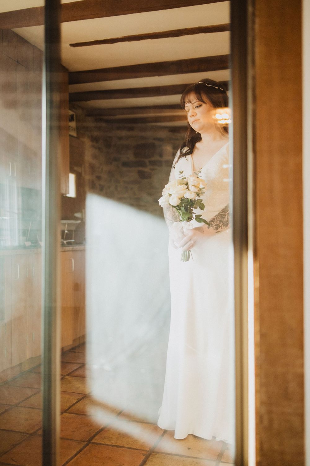 Bride in elegant white dress holding bouquet stands in sunlit rustic doorway.