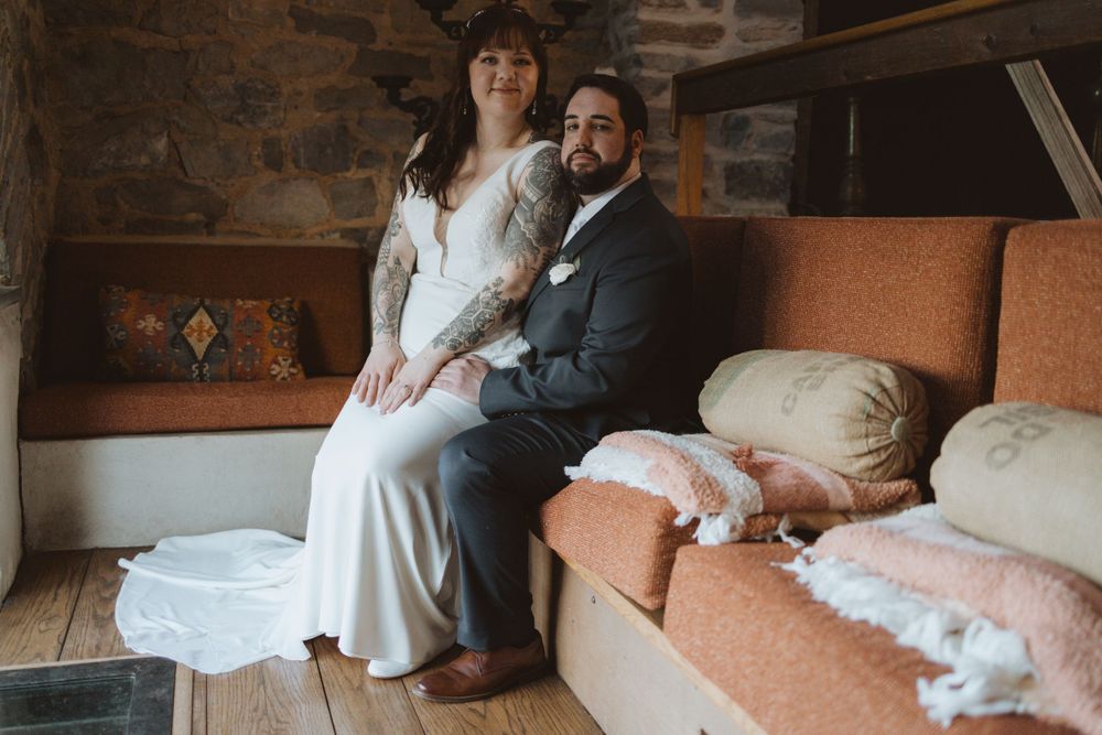Couple sits together on an orange couch in a rustic room with stone walls and white lace accents.