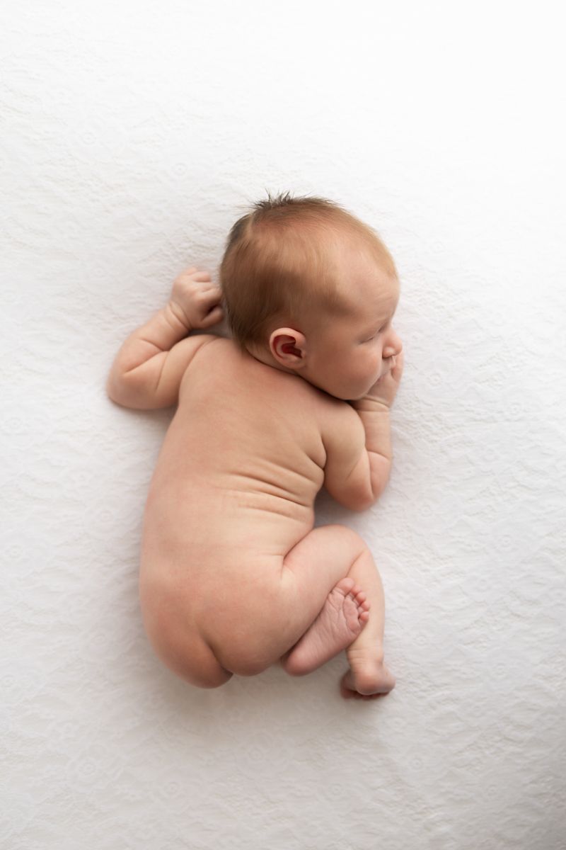 Newborn baby girl laying on white blanket, cuddled with legs and arms underneath her body, shot from above.