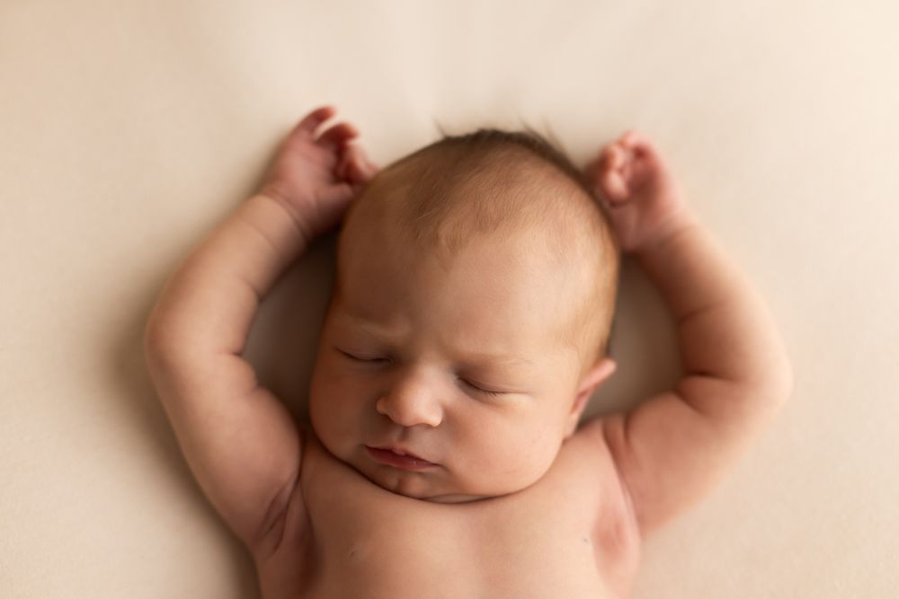 Newborn baby girl laying on off white blanket sleeping with her arms above her head.