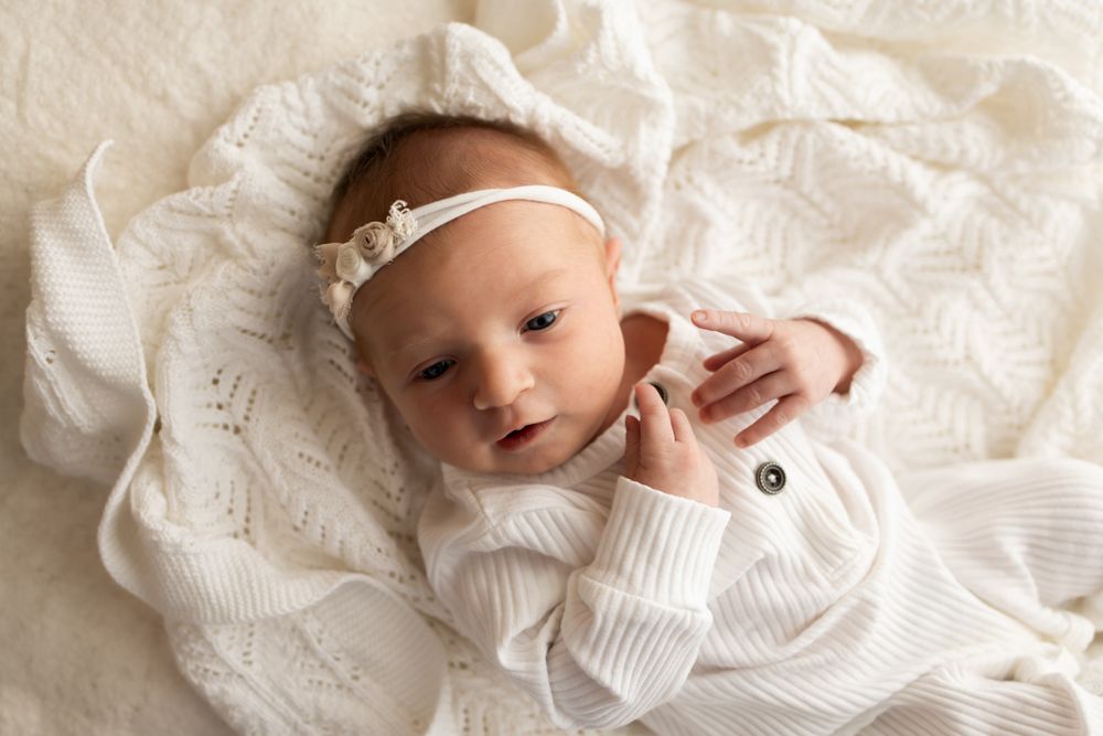 Newborn baby girl in white onesie laying on white crocheted blanket.