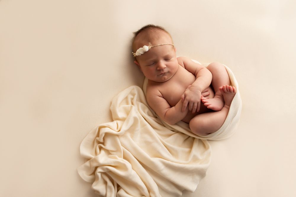 Newborn baby girl laying on a off white blanket, cuddled up with wrap around her and trailing off to the side.