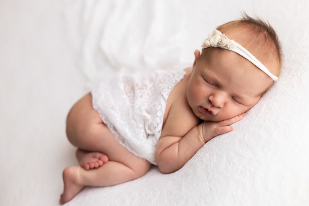 Newborn baby girl cuddled on hands and knees on a white blanket with white wrap over her back.
