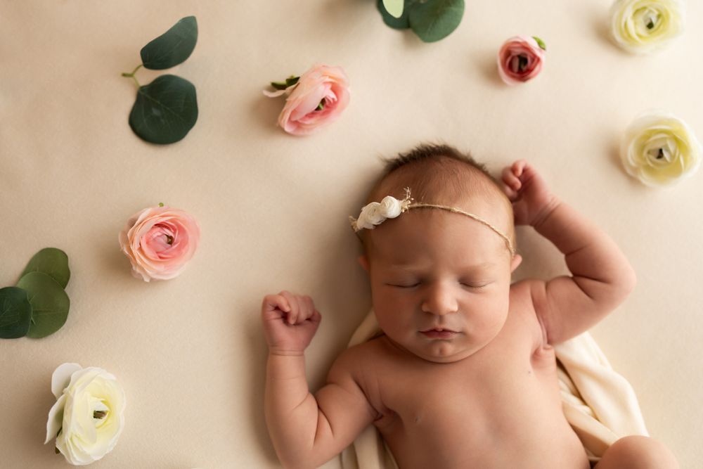 Newborn baby girl laying on a off white blanket with flowers surrounding her.